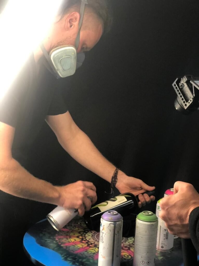 Gustavo Gomes, de máscara, pinta uma garrafa de vinho com spray