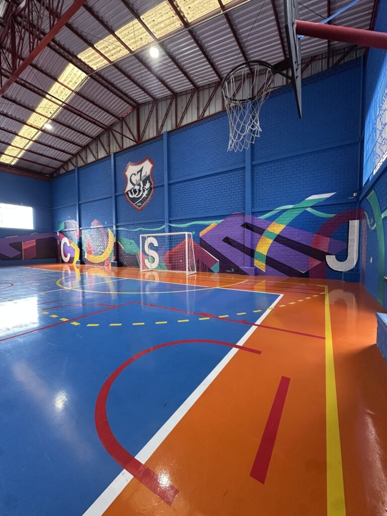 A quadra pintada em azul e laranja com as letras do colégio na parede ao fundo