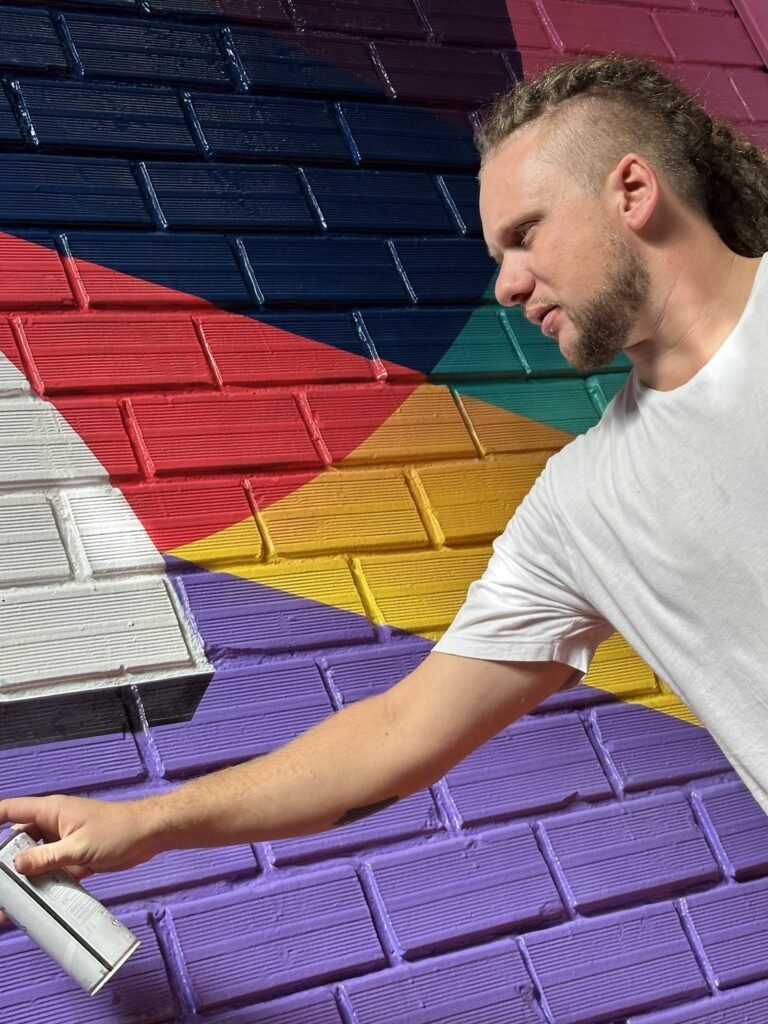 Gustavo Gomes pintando com spray os tijolos roxos e amarelos da parede