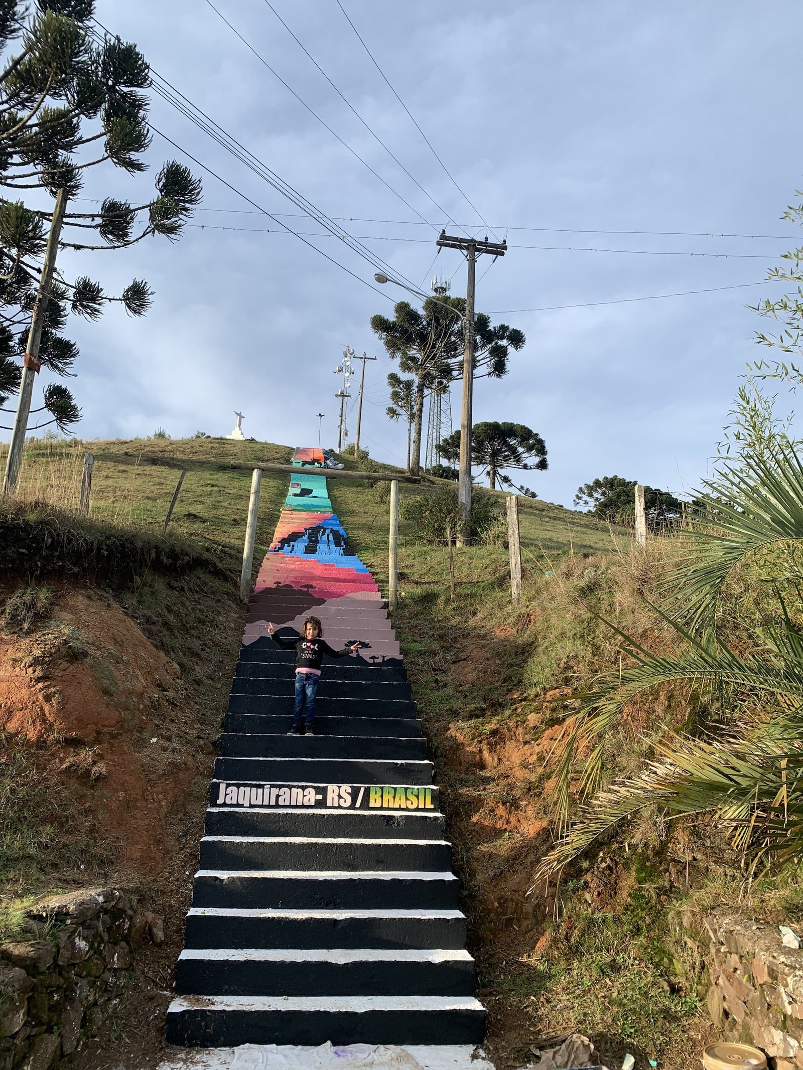 A escadaria colorida subindo o morro entre araucárias, com a inscrição Jaquirana RS Brasil nos degraus