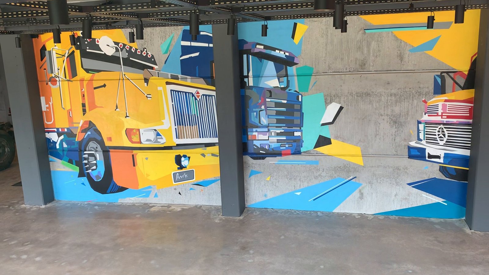 Mural com um caminhão amarelo e um vermelho em planos de cor, atrás das colunas do galpão