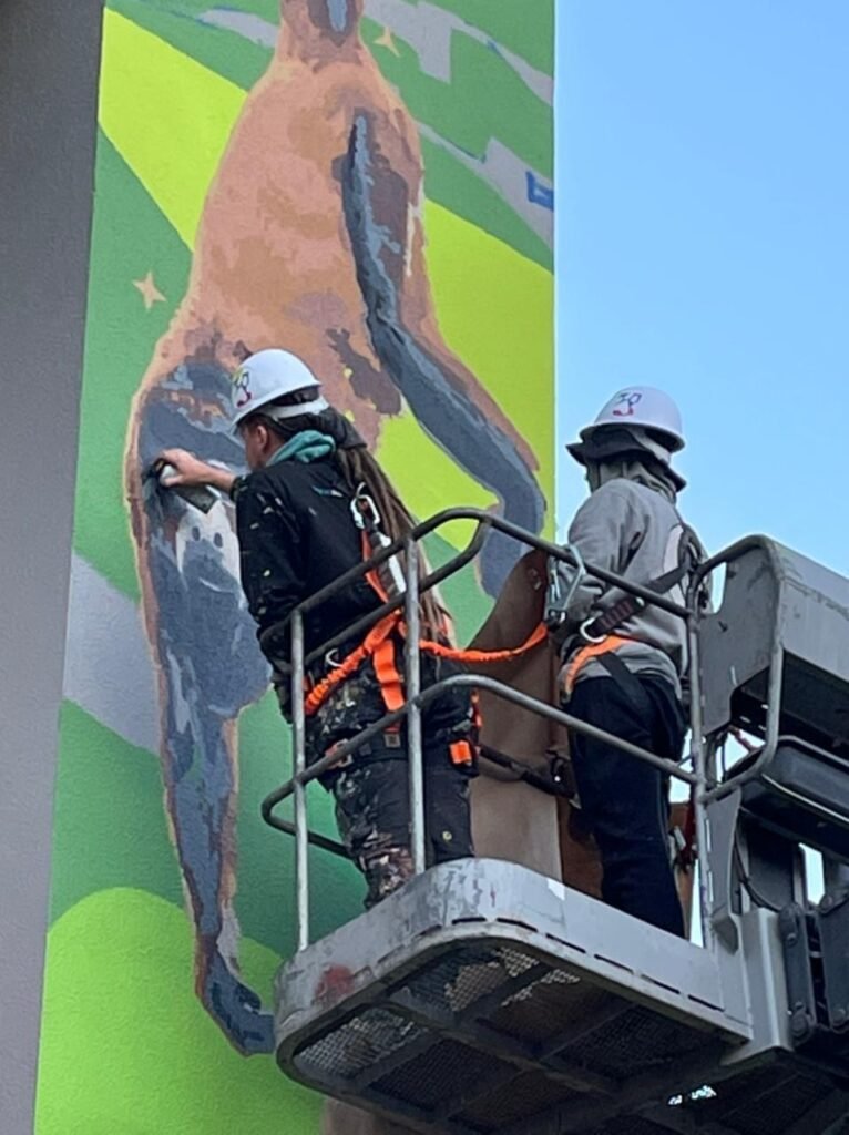 Duas pessoas de capacete pintando a figura de um bicho, de cima da plataforma elevatória