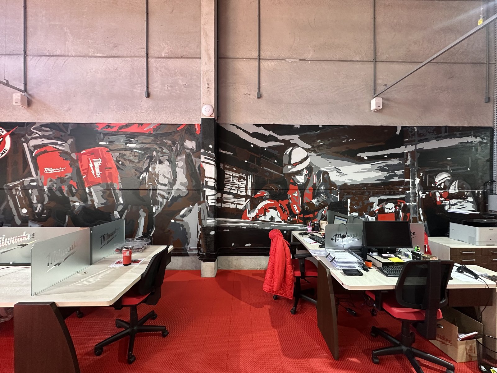 Escritório da Milwaukee com o mural em preto, branco e cinza atrás das mesas e o piso vermelho