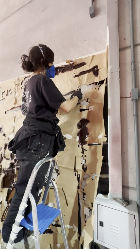 Integrante da equipe pinta de cima de uma escada, com as mesas cobertas de plástico
