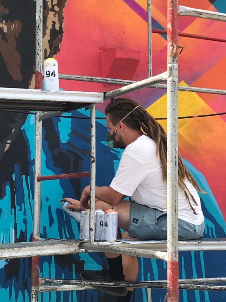 Pintor agachado no andaime, com as latas de spray ao lado, sobre o fundo laranja
