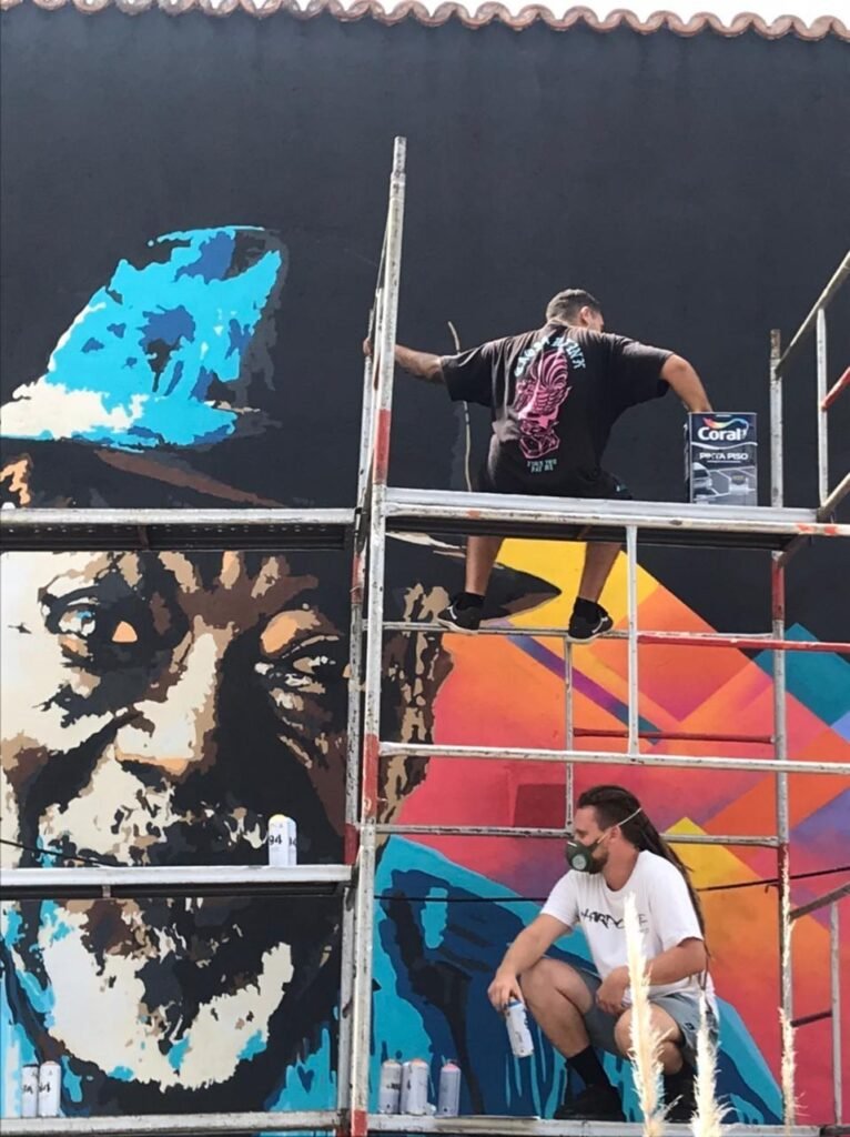 Duas pessoas pintando em alturas diferentes do andaime, na frente do homem de chapéu