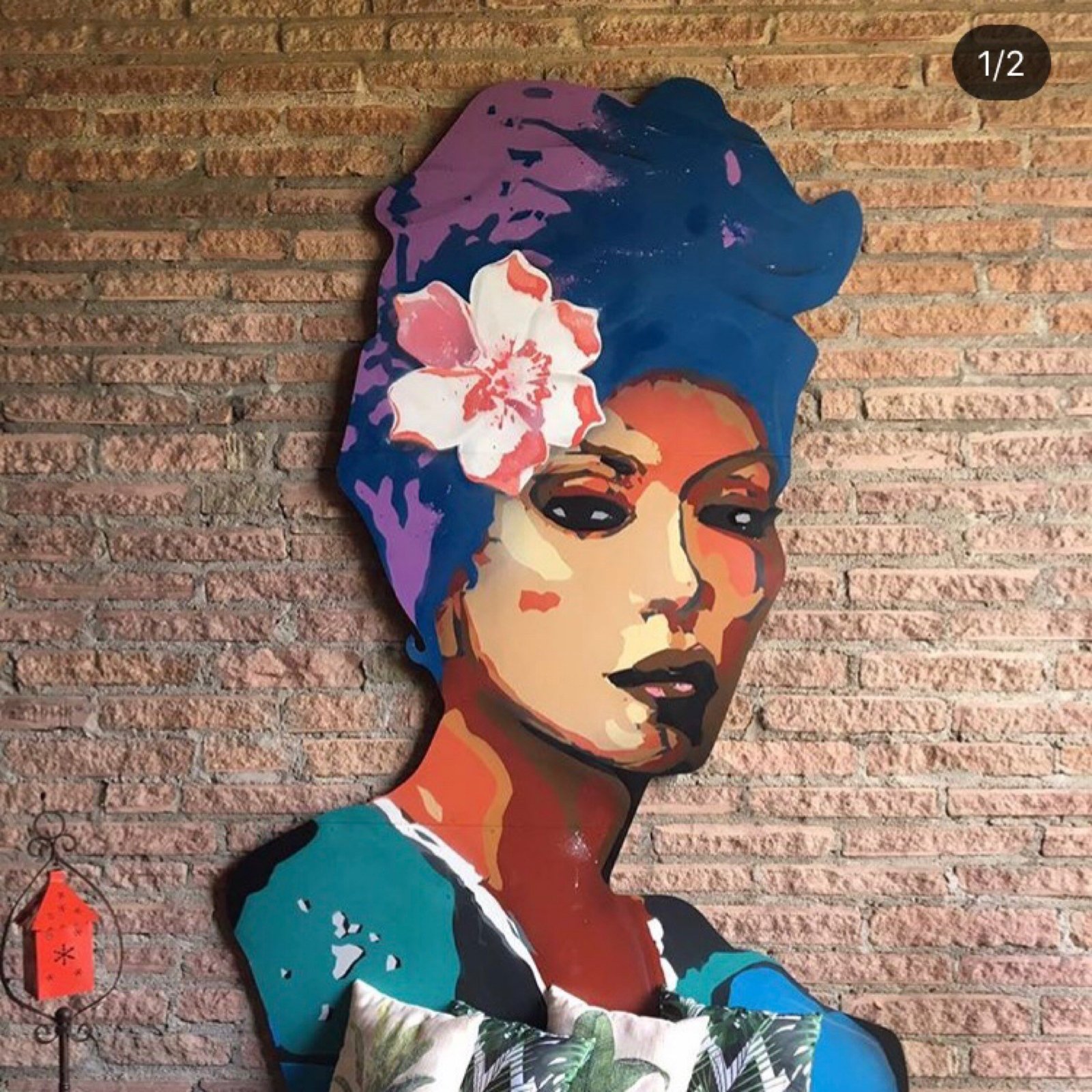 Figura recortada de uma mulher de lenço azul e flor no cabelo, encostada numa parede de tijolos