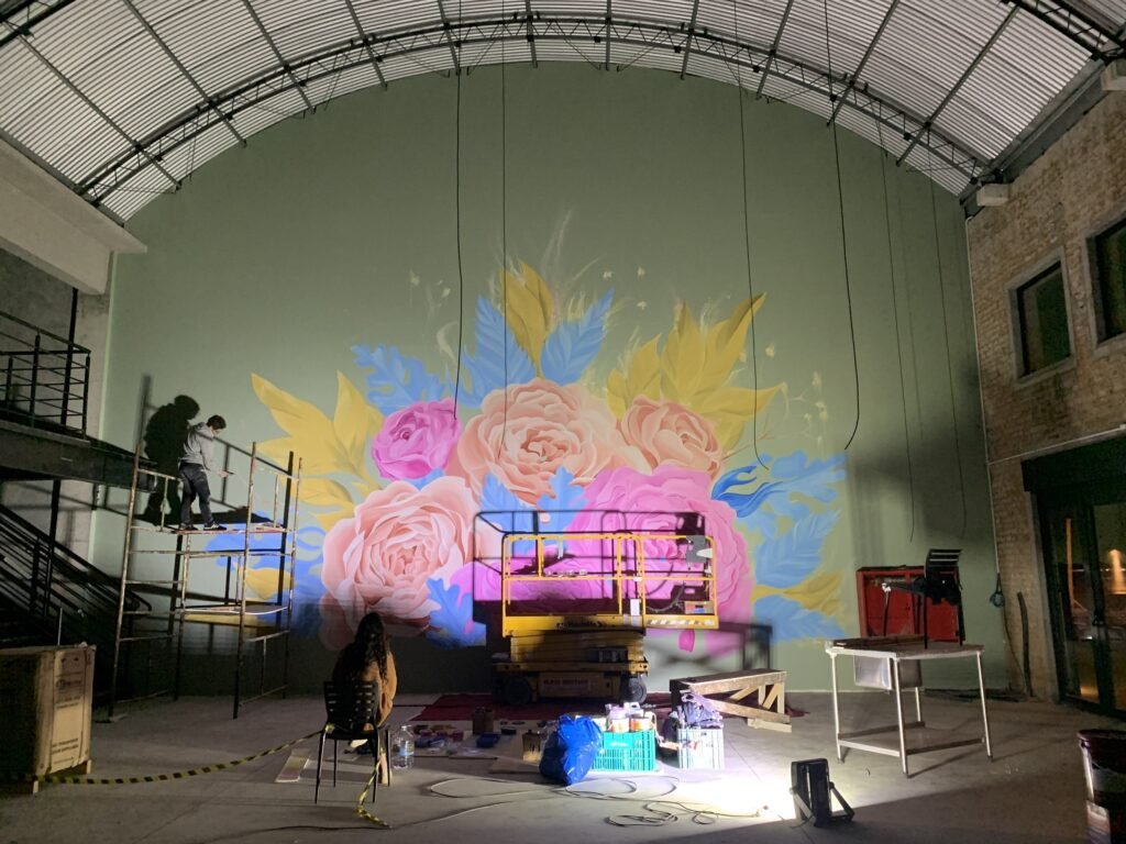 Galpão com o mural de rosas e folhas na parede do fundo, iluminado durante a pintura