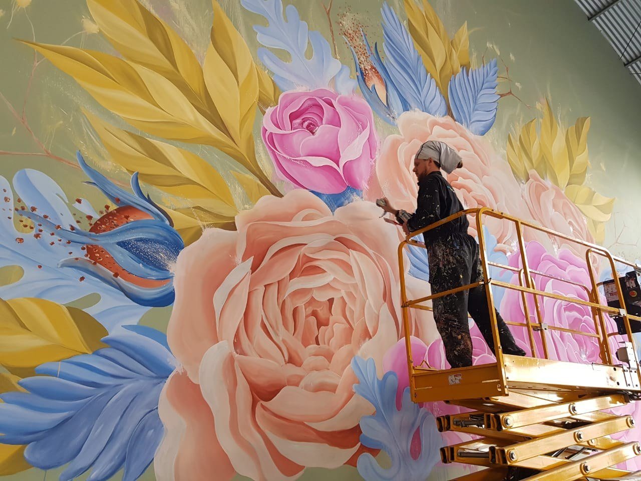 Rosas cor de pêssego e rosa entre folhas azuis e amarelas, com uma pessoa pintando na plataforma