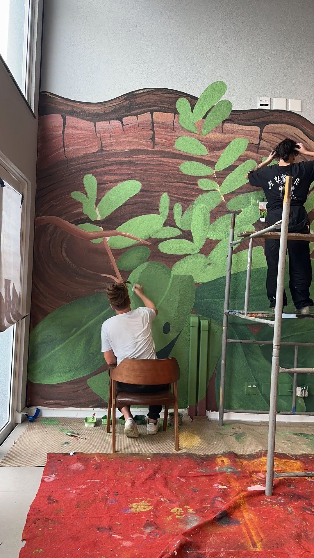 A parede inteira coberta de folhagem, com uma pessoa pintando e o piso protegido
