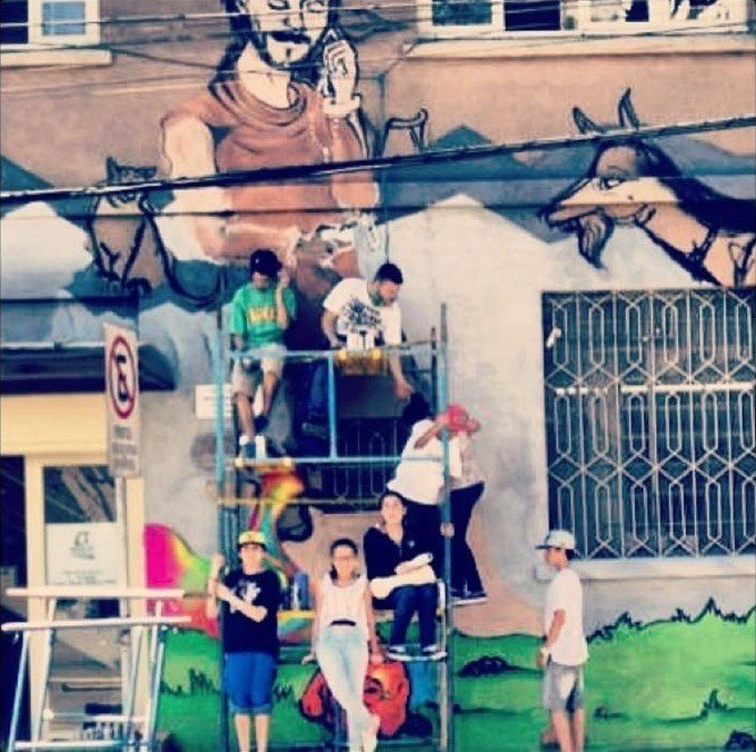 Mural pintado na fachada de um prédio, com andaime e crianças na calçada