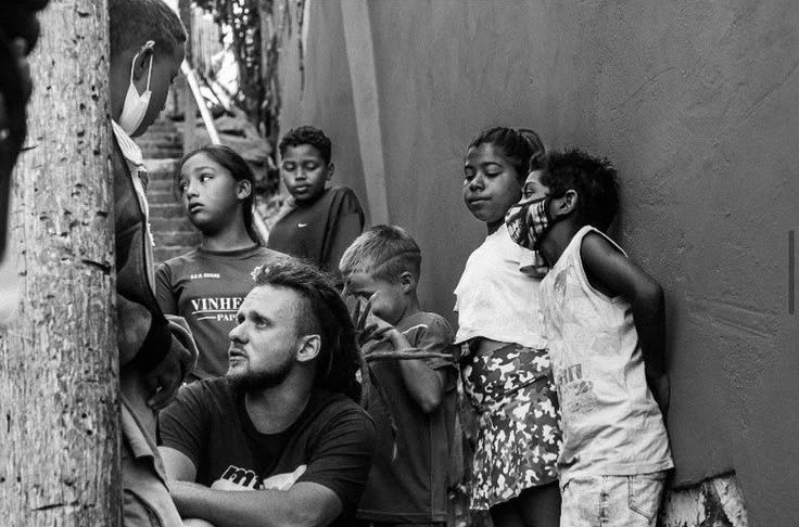 Foto em preto e branco de Gustavo Gomes agachado, conversando com crianças num beco