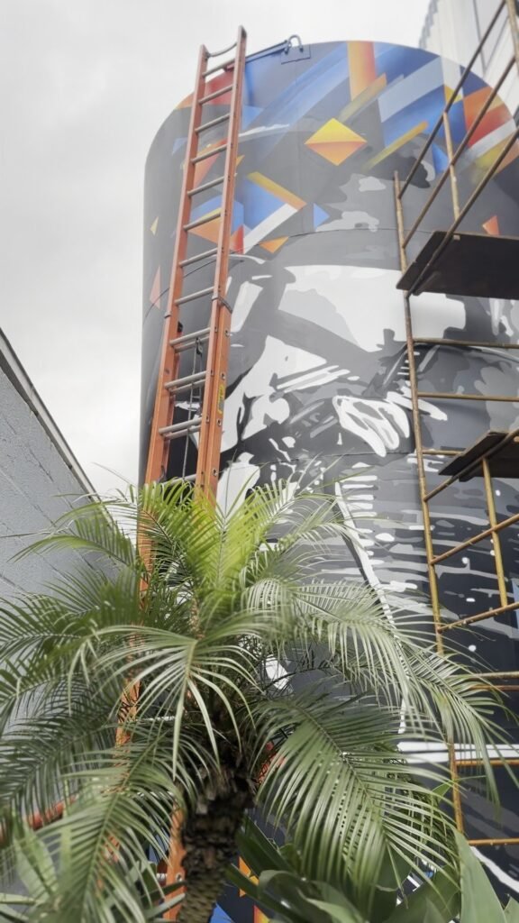 Trabalhador pintado em cinza num tanque cilíndrico, com andaime ao lado e uma palmeira na frente