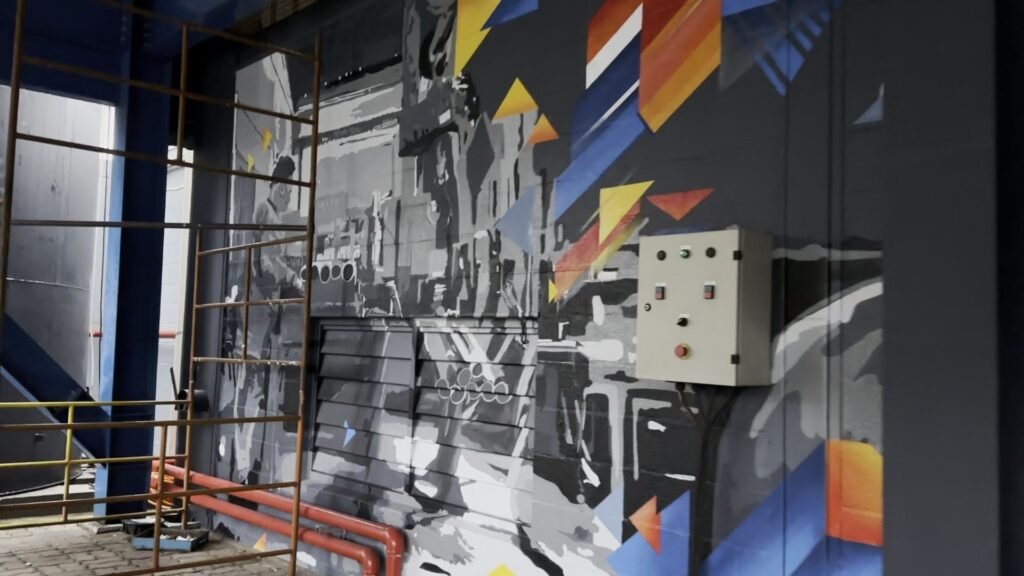 Mural de máquinas industriais em tons de cinza com triângulos azuis e laranja, dentro do galpão