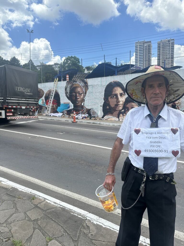 Um senhor de chapéu posa na calçada em frente ao mural