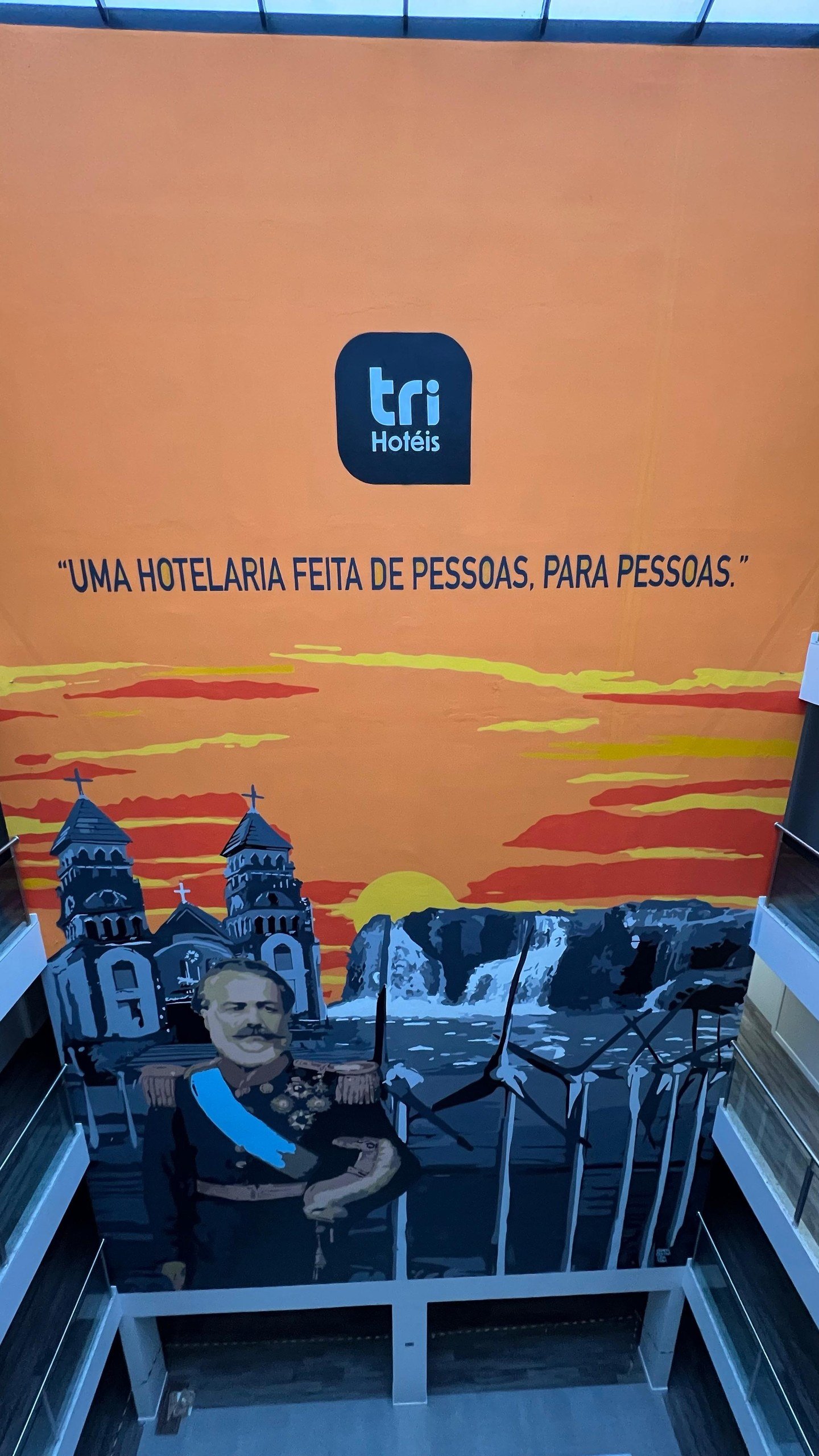 Mural de 17 metros no átrio do Tri Hotel Osório, com o pôr do sol laranja e a frase do hotel