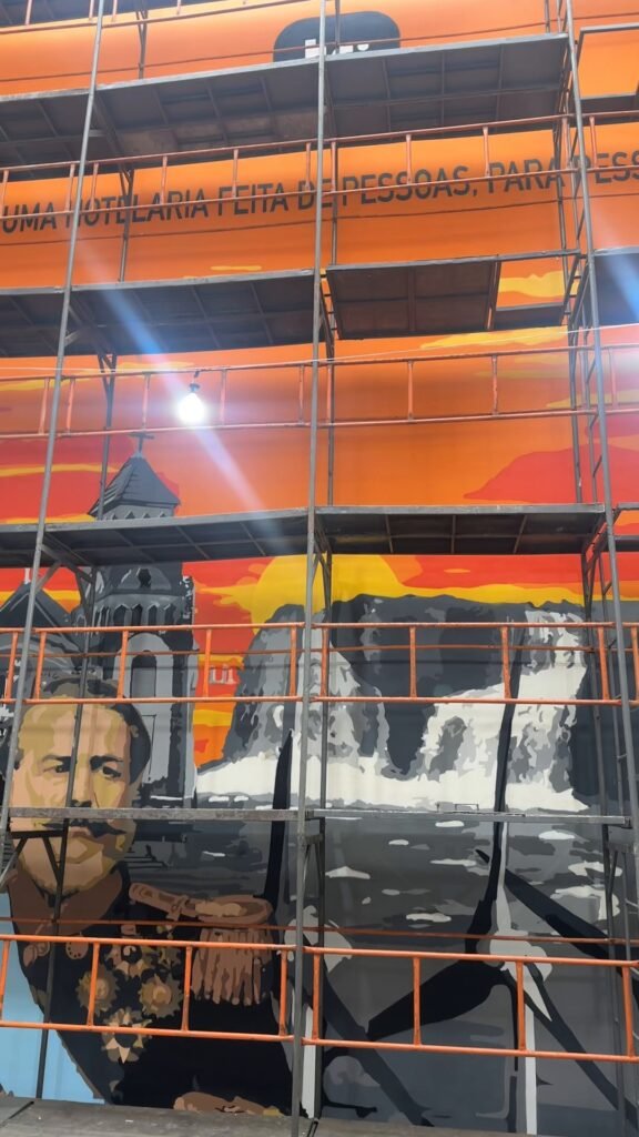 O retrato do General Osório sendo pintado atrás do andaime, com a cascata e o pôr do sol já prontos
