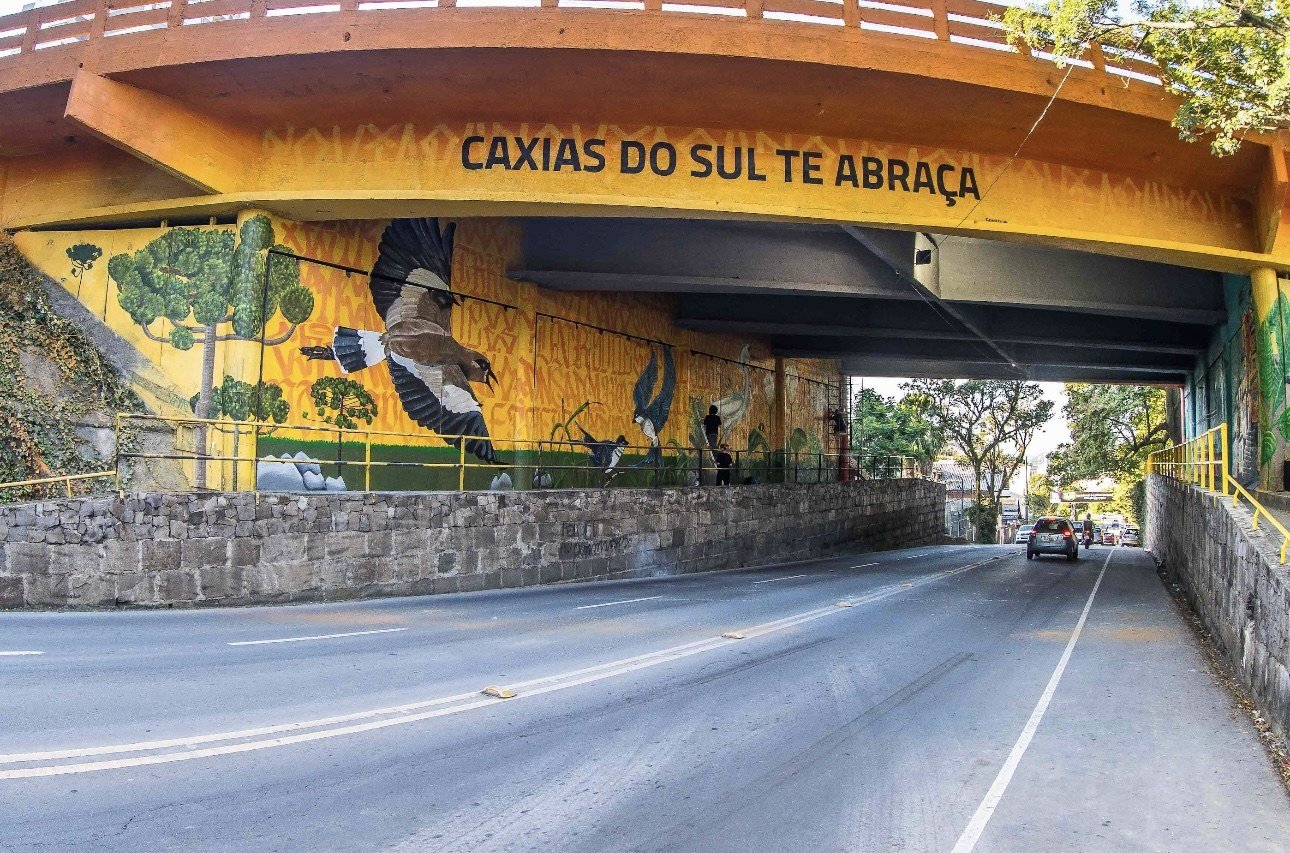 Viaduto pintado de amarelo com árvores, pássaros e a frase Caxias do Sul te abraça na viga